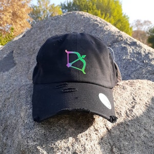 May include: Black distressed baseball cap with a colorful Sagittarius symbol. The cap features a curved brim and adjustable strap. The Sagittarius symbol is a bow and arrow design in shades of pink, green, and yellow.