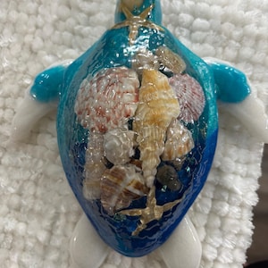 May include: A decorative sea turtle figurine featuring a glossy blue resin shell embedded with various natural seashells and two small starfish. The turtle has a smooth, light-colored head and flippers, with a gradient blue finish on the shell.