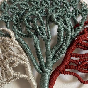 May include: A macrame wall hanging featuring a tree with three branches, each with a different color: beige, green, and red. The branches are intertwined and create a unique and intricate design.