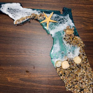 May include: A wall art piece shaped like the state of Florida, crafted from resin with a deep teal and green ocean gradient. It features white lacy wave effects, a gold starfish, a small sea turtle figure, and various seashells embedded in sand.