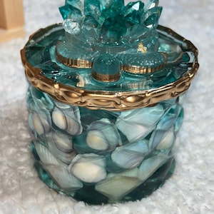 May include: A cylindrical container with a teal resin body embedded with numerous small white and light blue seashells. The lid features a sculpted teal crystal-like lotus flower topper and is finished with a decorative metallic gold rim.