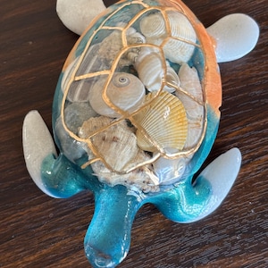 May include: A decorative turtle figurine with a shell filled with seashells and gold accents. The turtle's body is a mix of blue, orange, and clear resin, with white flipper-like appendages. The figurine is approximately 6 inches long.