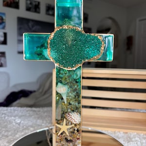 May include: A cross-shaped decorative object made of clear resin with a gold-toned metal frame. The interior contains green sand, small seashells, a starfish, and a central cluster of dark green, textured, crystalline mineral stones.