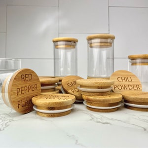 Puede incluir: Un juego de tarros de vidrio transparente con tapas de bambú. Las tapas están grabadas con varios nombres de especias, incluyendo "Red Pepper Flakes", "Garlic Powder", "Onion Powder" y "Chili Powder".