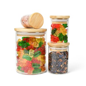 May include: Four glass jars with bamboo lids, filled with various items. The top jar is filled with pink Himalayan salt, the second jar is filled with colorful gummy bears, the third jar is filled with loose leaf tea, and the bottom jar is empty.