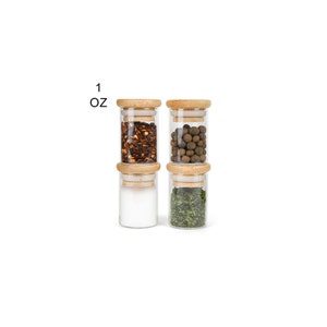 May include: Four glass spice jars with wooden lids, each filled with a different spice. The jars are arranged in two rows of two. The spices are red pepper flakes, black peppercorns, salt, and dried herbs. The jars are labeled "1 oz".