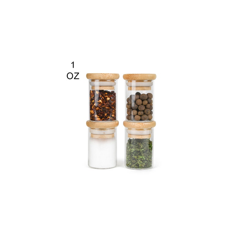 May include: Four glass spice jars with wooden lids, each filled with a different spice. The jars are arranged in two rows of two. The spices are red pepper flakes, black peppercorns, salt, and dried herbs. The jars are labeled "1 oz".