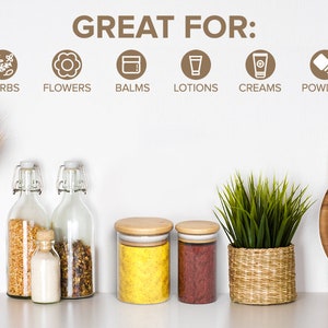 May include: Glass jars with wooden lids, filled with spices, herbs, and powders. The jars are on a white countertop with a wooden cutting board and a potted plant. The text "GREAT FOR:" is written on the wall above the jars, followed by a list of uses for the jars, including spices, herbs, flowers, balms, lotions, creams, powders, and more.