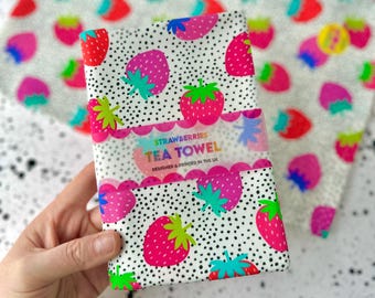 Può includere: Un asciugamano da tè bianco con un motivo a pois neri e una stampa di fragole colorate. L'asciugamano ha il testo "Strawberries Tea Towel" e "Designed & Printed in the UK".