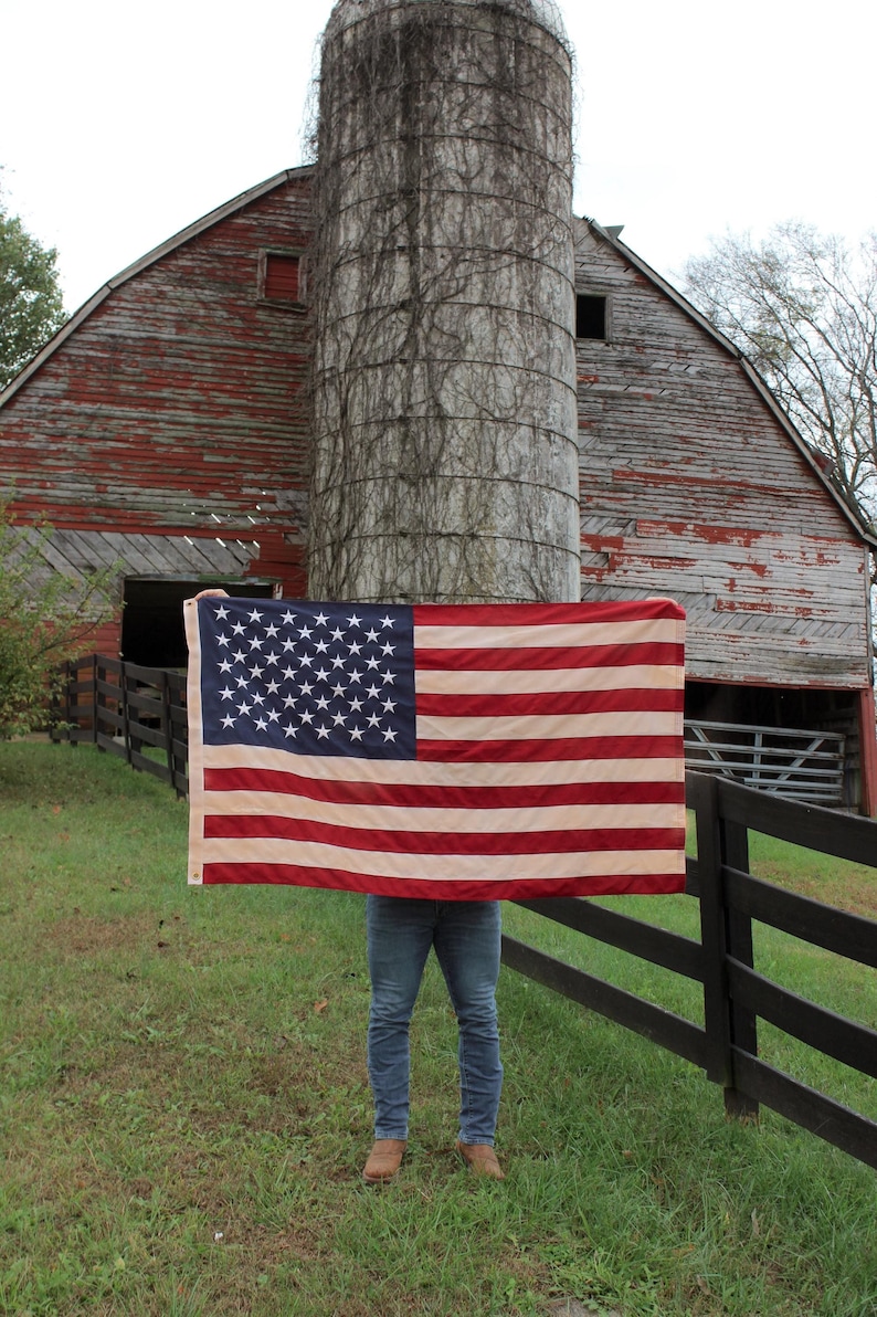 May include: A person holding a large American flag in front of a red barn with a silo. The flag is red, white, and blue with stars on the blue field. The barn is weathered and has a wooden fence in front of it.