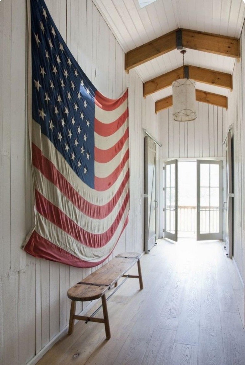 May include: A rustic wooden bench sits in a hallway with white walls and exposed wooden beams. A large American flag hangs on the wall behind the bench.
