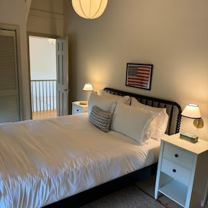 May include: A white bed with a black headboard and white bedding. There is a nightstand with two drawers on the right side of the bed. The room has a white ceiling with a round white lamp hanging from it. There is a framed picture of the American flag on the wall behind the bed.