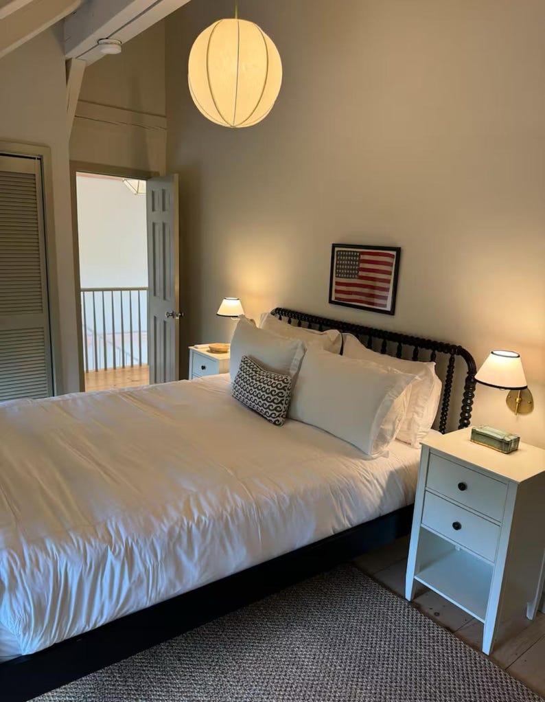 May include: A white bed with a black headboard and white bedding. There is a nightstand with two drawers on the right side of the bed. The room has a white ceiling with a round white lamp hanging from it. There is a framed picture of the American flag on the wall behind the bed.