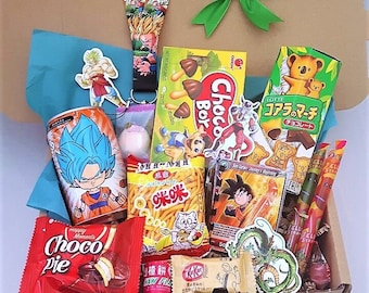 May include: A cardboard gift box containing an assortment of snacks and merchandise, including a Dragon Ball themed lanyard, a Choco Pie, a Choco Boy snack box, Lotte Koala's March cookies, a can with a blue-haired character, and various stickers.