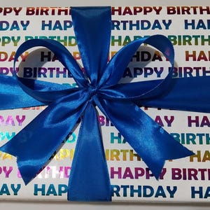 Może przedstawiać: Prostokątne pudełko prezentowe owinięte w biały papier z powtarzającym się napisem HAPPY BIRTHDAY w metalicznym tęczowym gradiencie. Pudełko jest przewiązane żywą niebieską satynową wstążką zawiązaną w dużą, ozdobną kokardę z wieloma pętelkami.