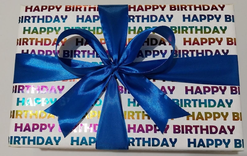 Może przedstawiać: Prostokątne pudełko prezentowe owinięte w biały papier z powtarzającym się napisem HAPPY BIRTHDAY w metalicznym tęczowym gradiencie. Pudełko jest przewiązane żywą niebieską satynową wstążką zawiązaną w dużą, ozdobną kokardę z wieloma pętelkami.