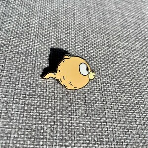 Puede incluir: Un pin de personaje de dibujos animados amarillo con un contorno marr&oacute;n. El personaje tiene un cuerpo redondo y una cola peque&ntilde;a. El personaje tiene un ojo grande y una boca peque&ntilde;a.