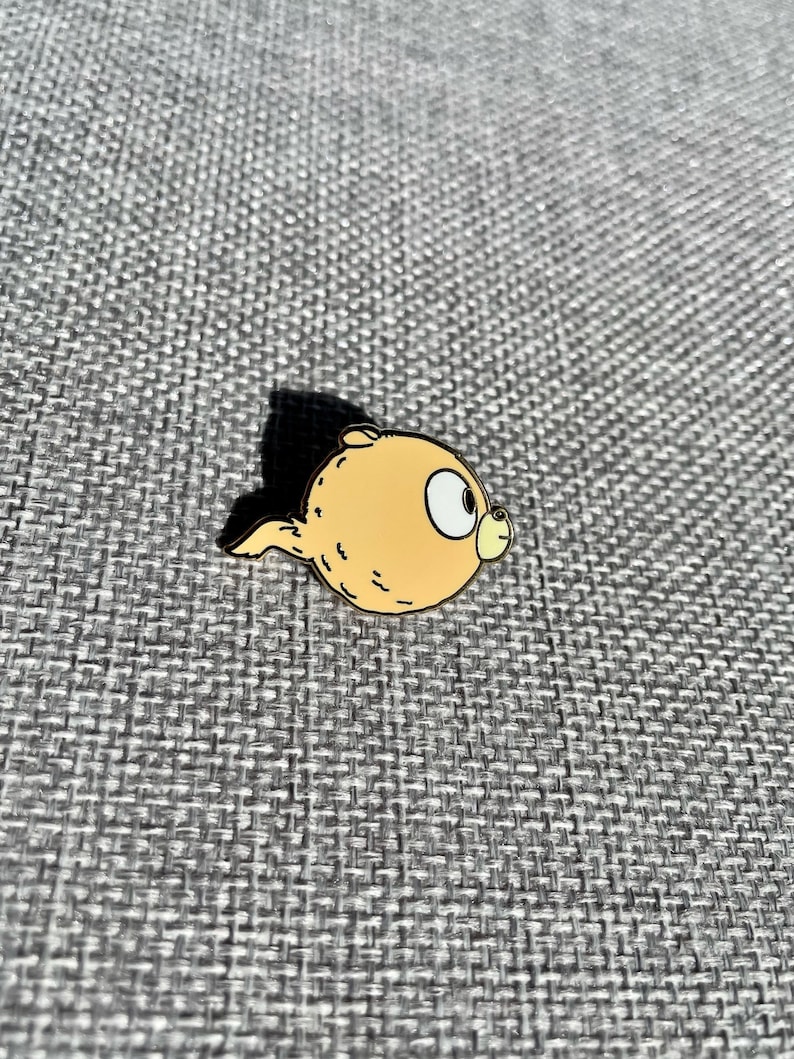 Puede incluir: Un pin de personaje de dibujos animados amarillo con un contorno marr&oacute;n. El personaje tiene un cuerpo redondo y una cola peque&ntilde;a. El personaje tiene un ojo grande y una boca peque&ntilde;a.