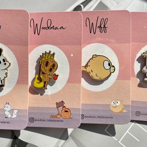 Puede incluir: Cuatro pines de esmalte con personajes de la serie de dibujos animados Hilda. Los pines est&aacute;n en tarjetas rosas y blancas con los nombres de los personajes "Twig", "Woodman", "Woff" y "Troll" impresos en las tarjetas. Los pines son de color dorado con contornos negros.