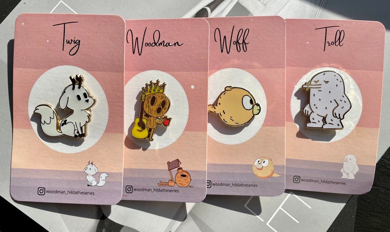 Puede incluir: Cuatro pines de esmalte con personajes de la serie de dibujos animados Hilda. Los pines est&aacute;n en tarjetas rosas y blancas con los nombres de los personajes "Twig", "Woodman", "Woff" y "Troll" impresos en las tarjetas. Los pines son de color dorado con contornos negros.