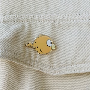 Puede incluir: Un pin de esmalte de color dorado con un pez de dibujos animados con un ojo grande y una cola peque&ntilde;a. El pez es de color marr&oacute;n claro con un ojo blanco y un contorno negro.