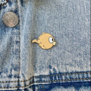 Puede incluir: Un pin de esmalte amarillo y naranja de un personaje de dibujos animados con cola. El personaje tiene un cuerpo redondo y un ojo grande y redondo. El pin est&aacute; sujeto a una chaqueta vaquera.