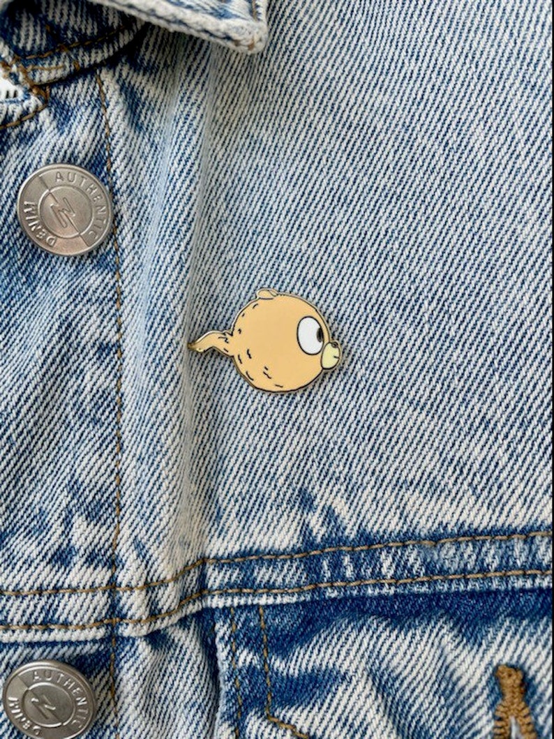 Puede incluir: Un pin de esmalte amarillo y naranja de un personaje de dibujos animados con cola. El personaje tiene un cuerpo redondo y un ojo grande y redondo. El pin est&aacute; sujeto a una chaqueta vaquera.