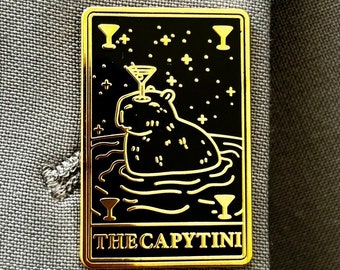 Puede incluir: Pin de esmalte dorado con fondo negro que representa a un capibara de dibujos animados con una copa de martini en la cabeza. El texto "THE CAPYTINI" está impreso debajo de la imagen.