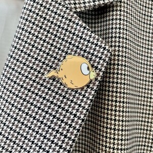 Puede incluir: Una chaqueta de punto de pata de gallo marr&oacute;n y blanco con un pin de esmalte dorado que representa un personaje de dibujos animados con un ojo grande y una cola.