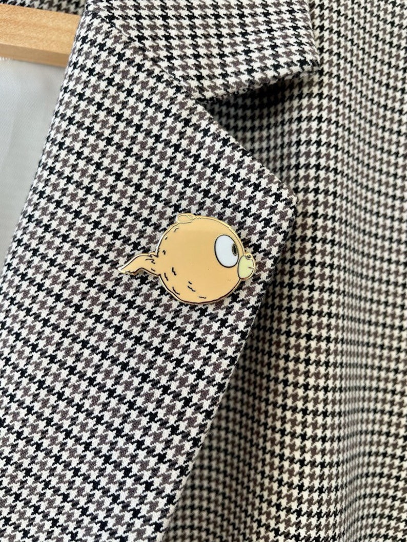 Puede incluir: Una chaqueta de punto de pata de gallo marr&oacute;n y blanco con un pin de esmalte dorado que representa un personaje de dibujos animados con un ojo grande y una cola.