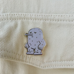 May include: A gray enamel pin featuring a cartoon monster with a grumpy expression. The pin has a gold border and is dotted with small black spots. The monster is standing with its arms at its sides. The pin is attached to a cream-colored denim jacket.
