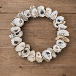 May include: A circular wreath made of oyster shells, showcasing a natural, coastal aesthetic. The shells are primarily white with subtle brown and gray markings, arranged on a dark brown, circular base. The wreath is set against a wooden background.