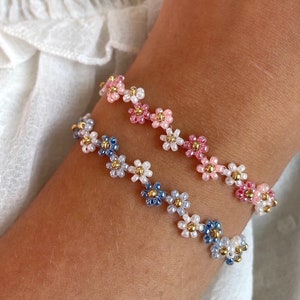 Könnte beinhalten: Zwei Perlenarmbänder mit Blumenmustern. Ein Armband ist rosa und weiß, das andere ist blau, weiß und gold.