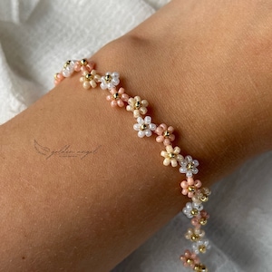 Op de afbeelding: Een delicate armband gemaakt van kleine, kleurrijke kralen in tinten roze, wit en goud. De kralen zijn gerangschikt in een patroon van kleine bloemetjes, wat een delicaat en vrouwelijk uiterlijk creëert.