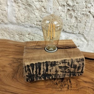 Puede incluir: Lámpara de mesa rústica de madera con una gran bombilla transparente de estilo Edison. La bombilla está colocada en una base de madera natural con un patrón oscuro y carbonizado en el borde. La lámpara está sobre una superficie de madera.