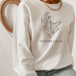 May include: White sweatshirt with a black line drawing of a woman holding a globe. The text "VENI VIDI AMA VI" is printed below the drawing.