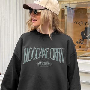 May include: A black sweatshirt with the text "BLOODAXE CREW" in a light blue font. Below the text is "THE ROAD OF BOND". The person in the image is wearing a tan baseball cap and sunglasses.