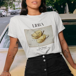 May include: White t-shirt featuring the word "LIBRA" above a vintage-style illustration of the Libra zodiac sign. The shirt also includes the text "The Charmer" and the dates "September 23rd - October 22nd".