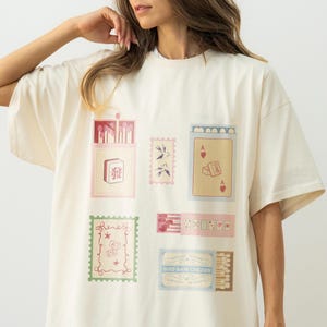 May include: An off-white short-sleeve t-shirt featuring a graphic print of various rectangular illustrations, including a mahjong tile, a playing card with an ace of hearts, postage stamps, and matchboxes with detailed line art and soft colors.