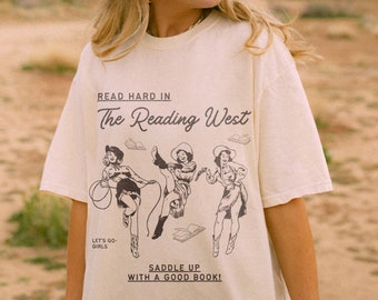 May include: A white t-shirt with a graphic print of three women riding horses with books. The text reads "Read hard in The Reading West, Let's go-girls, Saddle up with a good book!"