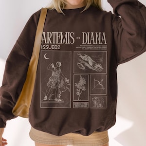 May include: A dark brown sweatshirt with the text "ARTEMIS - DIANA ISSUE02" in white. The sweatshirt features a graphic design with illustrations of a woman, animals, and a bow and arrow. The design is in a vintage style.