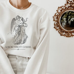 May include: White sweatshirt with a black and white illustration of two figures embracing. The text "AMOR VINCIT OMNIA" is printed below the illustration.