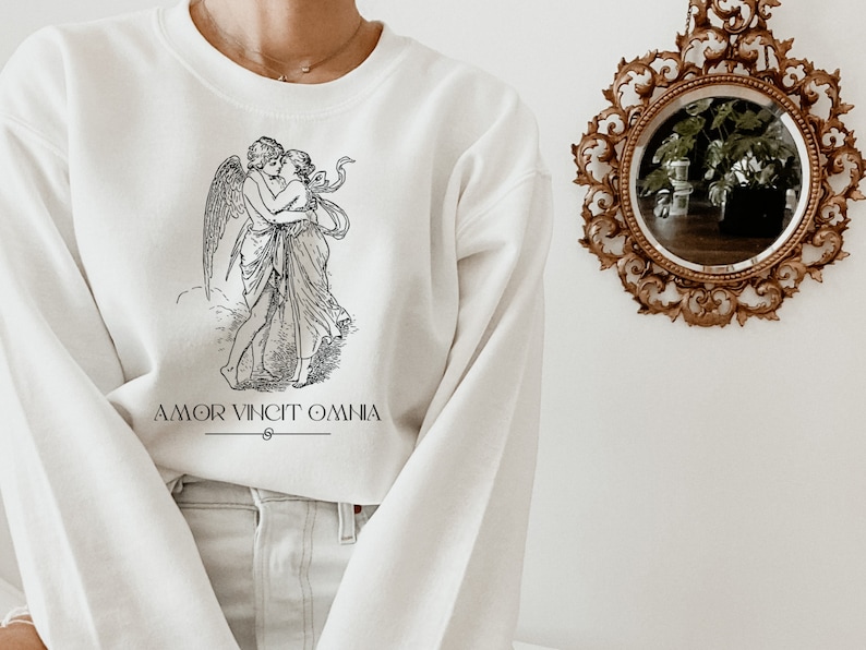 May include: White sweatshirt with a black and white illustration of two figures embracing. The text "AMOR VINCIT OMNIA" is printed below the illustration.