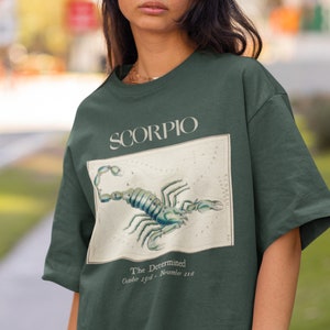 Op de afbeelding: Een donkergroen T-shirt met een vintage illustratie van een schorpioen en de tekst "SCORPIO The Determined October 23rd - November 21st"