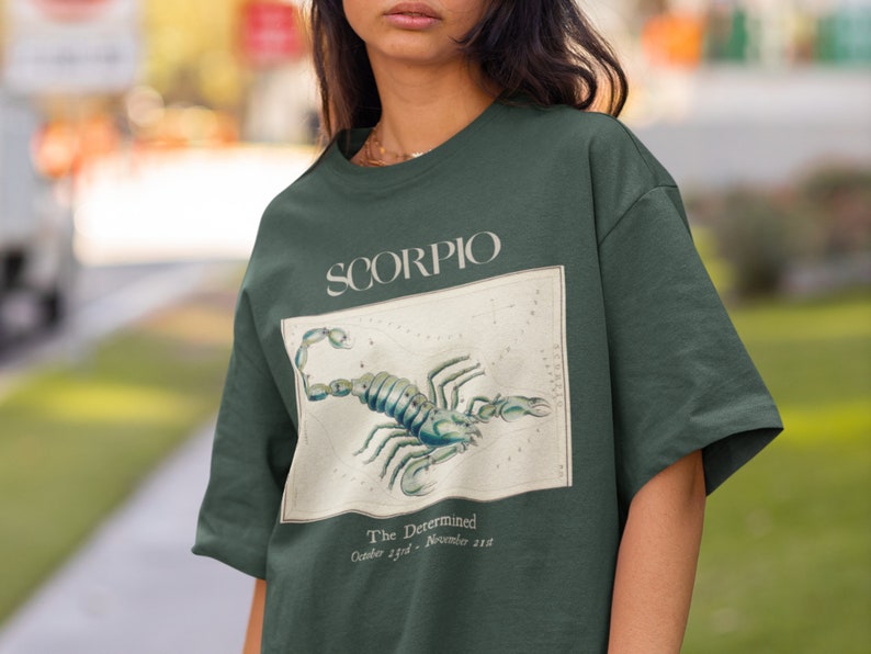 Op de afbeelding: Een donkergroen T-shirt met een vintage illustratie van een schorpioen en de tekst "SCORPIO The Determined October 23rd - November 21st"
