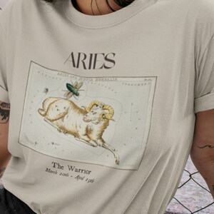 Pu&ograve; includere: Una t-shirt beige con un'illustrazione vintage della costellazione dell'Ariete, un ariete e una mosca. Il testo "Aries" &egrave; stampato in nero nella parte superiore dell'illustrazione. Il testo "The Warrior" e "March 20th - April 19th" &egrave; stampato sotto l'illustrazione.