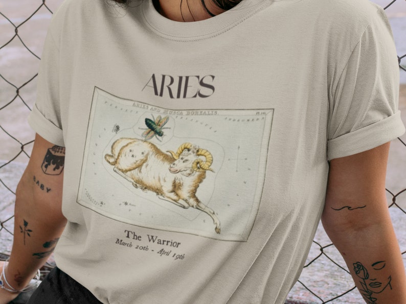 Pu&ograve; includere: Una t-shirt beige con un'illustrazione vintage della costellazione dell'Ariete, un ariete e una mosca. Il testo "Aries" &egrave; stampato in nero nella parte superiore dell'illustrazione. Il testo "The Warrior" e "March 20th - April 19th" &egrave; stampato sotto l'illustrazione.