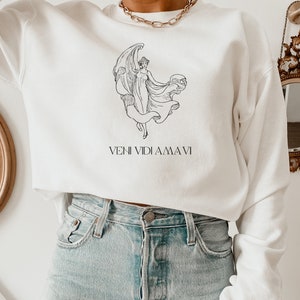 May include: White cropped sweatshirt with a black line drawing of a woman with wings and a globe. The text "VENI VIDI AMA VI" is printed below the drawing.