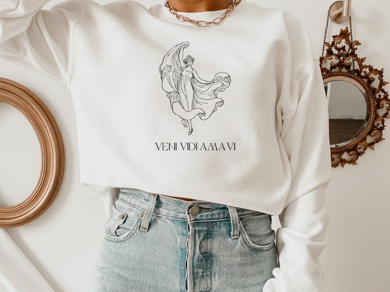May include: White cropped sweatshirt with a black line drawing of a woman with wings and a globe. The text "VENI VIDI AMA VI" is printed below the drawing.