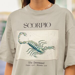 Op de afbeelding: Een lichtgrijs T-shirt met een vintage illustratie van het sterrenbeeld Schorpioen en de tekst "SCORPIO" en "The Determined October 23rd - November 21st".
